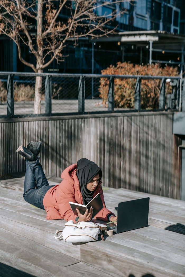 Young focused Muslim female student lying on wooden roof using laptop while preparing for exam in sunny autumn day