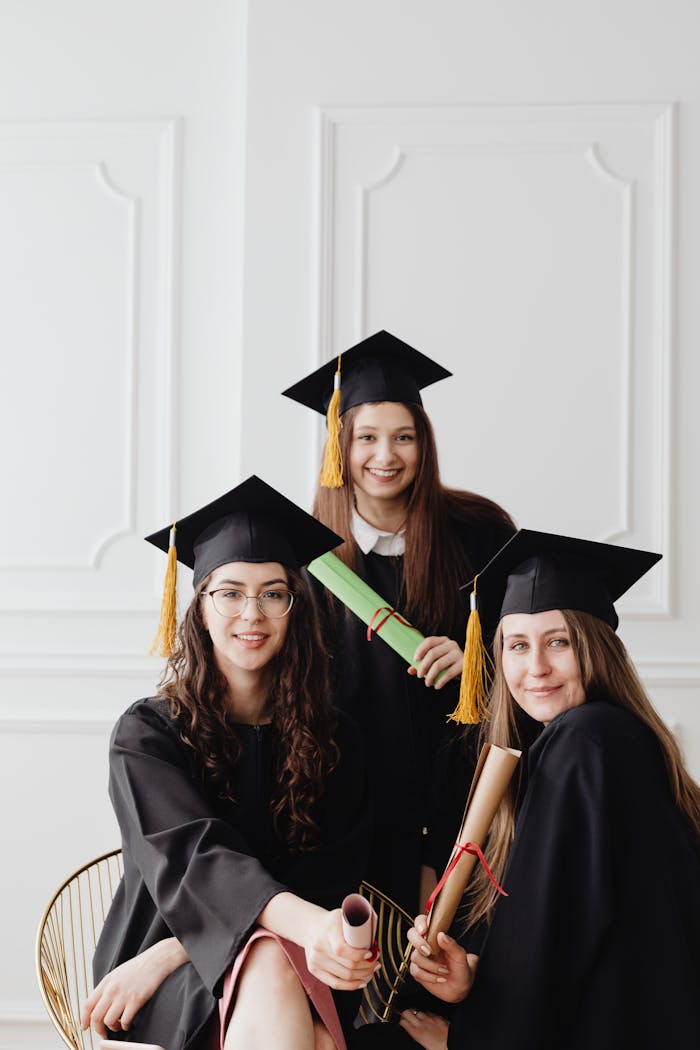 Three female graduates in caps and gowns posing with diplomas indoors.